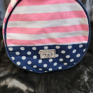 May include: A pink and white striped duffel bag with a blue polka dot bottom. The bag has a navy blue handle and a brown leather patch with the text "HILFIGER 1985" on it.