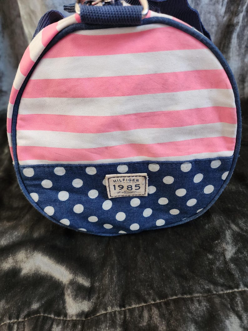 May include: A pink and white striped duffel bag with a blue polka dot bottom. The bag has a navy blue handle and a brown leather patch with the text "HILFIGER 1985" on it.