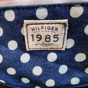 May include: A close-up of a blue and white polka dot fabric with a brown rectangular tag that reads "HILFIGER STATE OF NEW YORK 1985 Tommy Hilfiger DENIM".