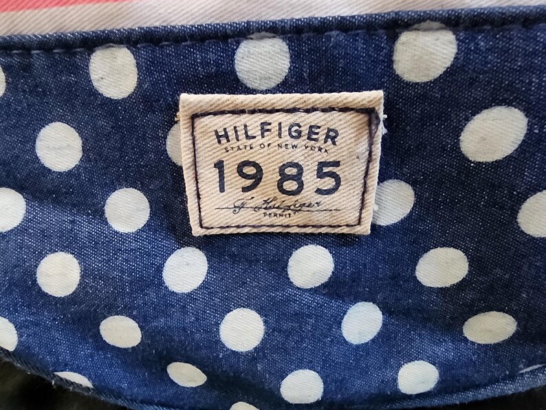 May include: A close-up of a blue and white polka dot fabric with a brown rectangular tag that reads "HILFIGER STATE OF NEW YORK 1985 Tommy Hilfiger DENIM".