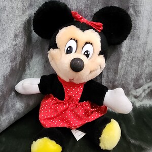 Puede incluir: Un juguete de peluche de Minnie Mouse con un vestido rojo con lunares blancos y un lazo rojo. El juguete tiene orejas negras, una nariz negra y zapatos amarillos.