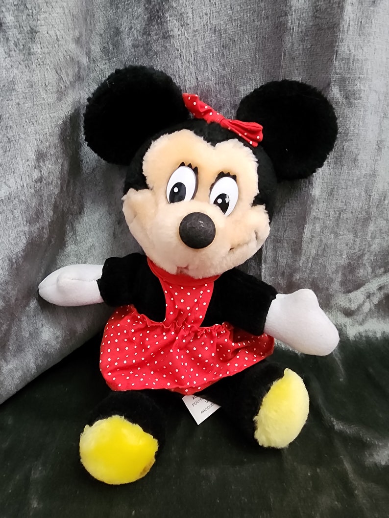 Puede incluir: Un juguete de peluche de Minnie Mouse con un vestido rojo con lunares blancos y un lazo rojo. El juguete tiene orejas negras, una nariz negra y zapatos amarillos.