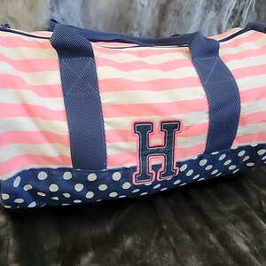 May include: A pink and white striped duffel bag with blue polka dots and a blue handle. The bag has a large embroidered letter "H" on the front.
