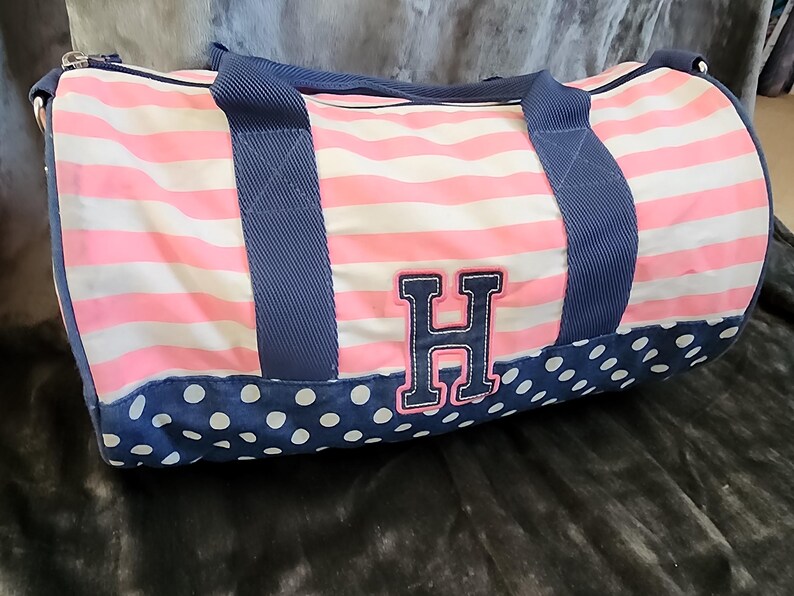 May include: A pink and white striped duffel bag with blue polka dots and a blue handle. The bag has a large embroidered letter "H" on the front.