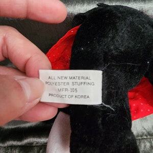 Puede incluir: Un juguete de peluche negro y rojo con un pico amarillo y patas blancas. El juguete tiene una etiqueta que dice "ALL NEW MATERIAL POLYESTER STUFFING MFR-105 PRODUCT OF KOREA".