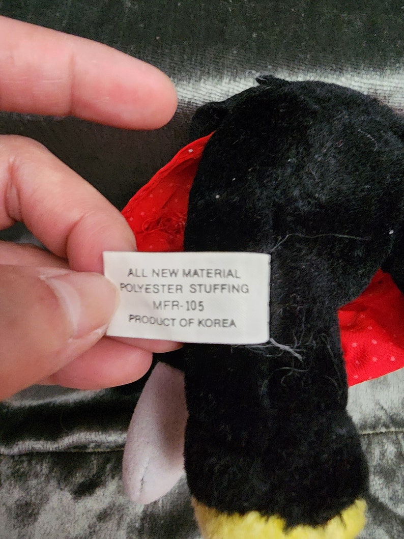 Puede incluir: Un juguete de peluche negro y rojo con un pico amarillo y patas blancas. El juguete tiene una etiqueta que dice "ALL NEW MATERIAL POLYESTER STUFFING MFR-105 PRODUCT OF KOREA".