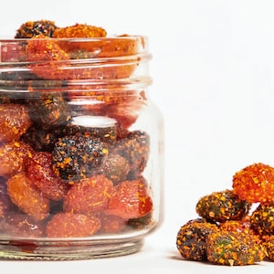 May include: A glass jar filled with red, orange, and black gummy candies. The candies are covered in a sugary coating. Some candies are scattered on a white surface.