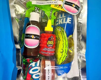 May include: A clear plastic bag filled with a variety of snacks, including a green pickle, a small bottle of hot sauce, a small jar of red powder, a green and red bag of chips, and a small bottle of brown liquid. The bag is labeled "Chammoy Pickle Kit" and includes a sticker that says "Miami Pika".