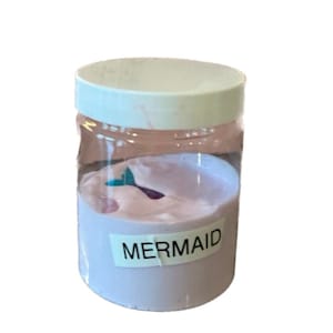 Puede incluir: Un tarro de plástico transparente con tapa blanca lleno de slime rosa claro. El tarro tiene una etiqueta que dice "MERMAID".