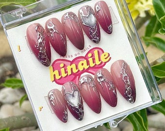 Puede incluir: Un conjunto de diez uñas postizas con un diseño degradado en tonos rosa y rojo. Cada uña presenta un corazón plateado y detalles plateados intrincados. Las uñas se muestran en una caja de plástico transparente con el texto "hinaile" y "press on nails".