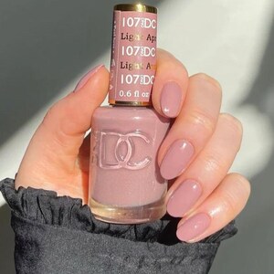 Pode incluir: Um frasco de verniz de unhas de damasco claro e unhas pintadas a condizer. O frasco tem um tom rosa-malva com o texto "107DC Light Apricot" e "0,6 fl oz" impresso no rótulo. As unhas são ovais e brilhantes.