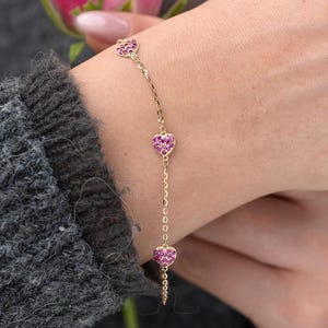 Op de afbeelding: Een gouden kettingarmband met drie roze hartvormige bedels. Elke bedel is bedekt met kleine roze steentjes.