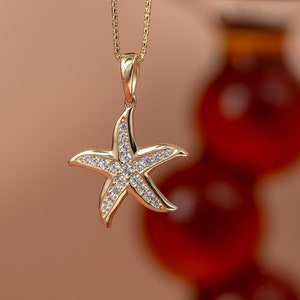 May include: A gold necklace with a starfish pendant encrusted with clear cubic zirconia stones. The necklace is hanging against a blurred background of red spheres.
