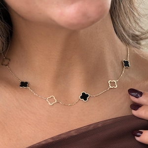 May include: A gold necklace with clover-shaped pendants in black and white. The necklace is worn around the neck. The person is wearing a brown strapless top. The necklace is a delicate piece of jewellery.