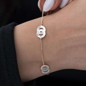 Puede incluir: Una delicada pulsera dorada con dos colgantes circulares entrelazados y un tercer colgante al final. Los colgantes tienen un centro blanco nacarado. La pulsera se lleva en una muñeca con una manga negra.