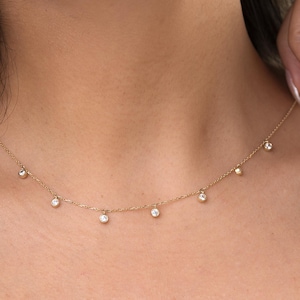 May include: A delicate gold necklace featuring multiple small, clear gemstones. The necklace is worn around the neck, with the gemstones evenly spaced along the chain. The jewelry is simple and elegant.