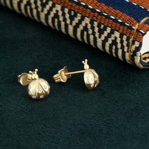 May include: A pair of gold ladybug earrings with clear sparkling stones. The earrings are on a dark green surface.