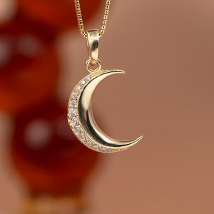 Può includere: Una collana con un ciondolo a forma di mezzaluna d'oro, decorata con pietre trasparenti scintillanti sul bordo. La collana è su una catena delicata.