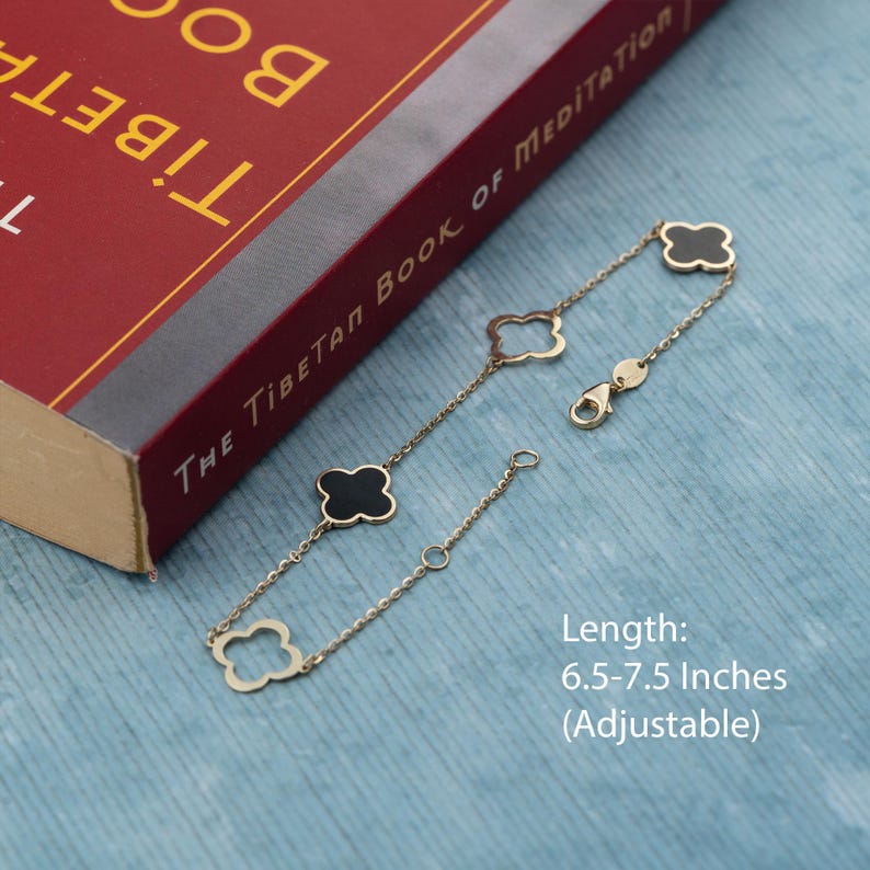 May include: Gold-toned bracelet featuring clover-shaped charms with black enamel centers. The adjustable bracelet, with a length of 6.5-7.5 inches, is displayed on a blue surface next to a red book titled "The Tibetan Book of Meditation."