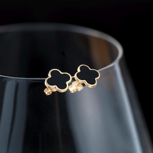 May include: A pair of gold-coloured stud earrings, each with a four-leaf clover design and a black centre. The earrings are set against a blurred background, highlighting their elegant shape and colour.