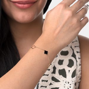 May include: A gold-colored bracelet featuring a black clover-shaped charm and several open diamond-shaped links. The delicate chain is worn on a wrist, showcasing the jewelry's design and color against the skin.