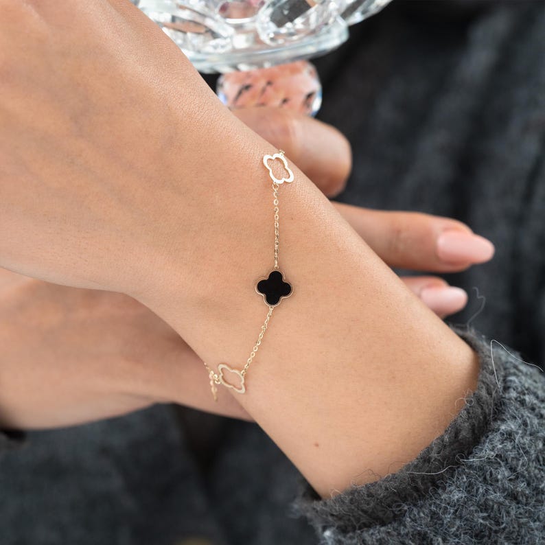 May include: A gold-colored bracelet featuring a clover-shaped charm with a black center, connected by a delicate chain to two openwork clover designs. The bracelet is worn on a wrist against a gray background.