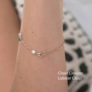 May include: Gold-toned chain bracelet with a lobster clasp closure. The bracelet features small, dark-colored rectangular charms and a delicate chain design. The image shows the bracelet on a person's arm.