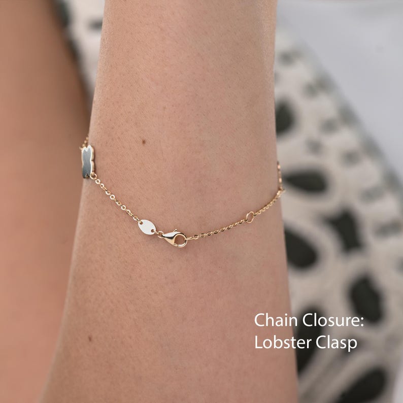 May include: Gold-toned chain bracelet with a lobster clasp closure. The bracelet features small, dark-colored rectangular charms and a delicate chain design. The image shows the bracelet on a person's arm.