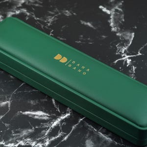 May include: A rectangular, emerald green jewelry box with a gold logo that reads "DD / DANA / DANO". The box is closed and sits on a black and white marble surface. The box is likely for jewelry or other small accessories.