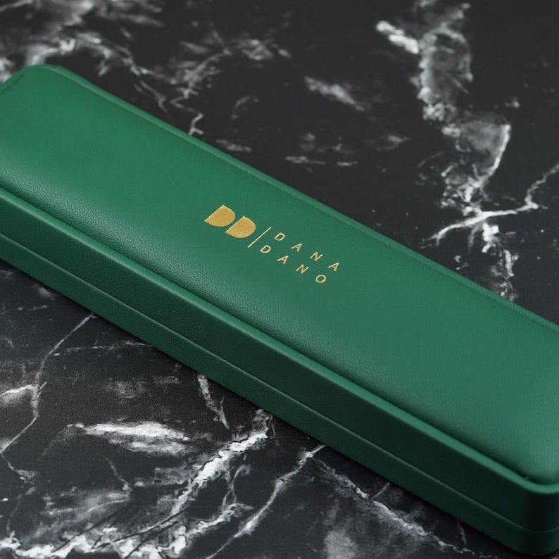 May include: A rectangular, emerald green jewelry box with a gold logo that reads "DD / DANA / DANO". The box is closed and sits on a black and white marble surface. The box is likely for jewelry or other small accessories.