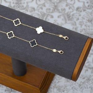 May include: Two gold-colored clover charm bracelets displayed on a gray jewelry stand. One bracelet features white clover charms, the other has black clover charms. The bracelets are fastened with a lobster clasp.