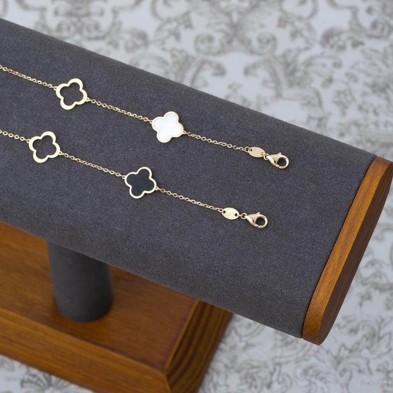 May include: Two gold-colored clover charm bracelets displayed on a gray jewelry stand. One bracelet features white clover charms, the other has black clover charms. The bracelets are fastened with a lobster clasp.