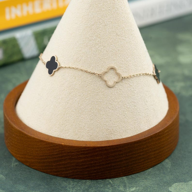 May include: A gold-toned bracelet with clover-shaped charms, some filled with black enamel. The bracelet is displayed on a white cone-shaped stand with a wooden base. The background includes a book and a green surface.