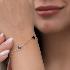May include: A gold bracelet featuring three black clover-shaped charms. The bracelet has a delicate chain. The charms are evenly spaced, creating a simple and elegant design. The bracelet is worn on a wrist.
