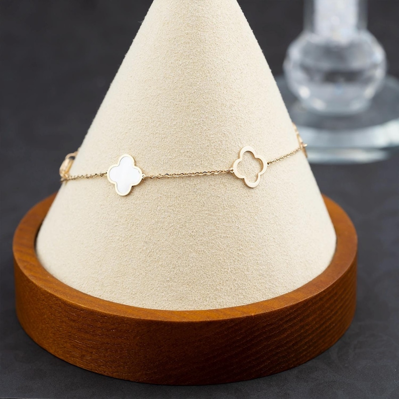 Puede incluir: Un brazalete de cadena dorada con tres dijes de tr&eacute;bol blanco. El brazalete se muestra en un soporte de madera con una parte superior de tela crema.