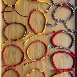 Pode incluir: Uma coleção de pulseiras artesanais em várias cores, incluindo vermelho, amarelo e multicolorido. As pulseiras são feitas de cordão tecido e possuem fechos ajustáveis. São exibidas em uma superfície de madeira.