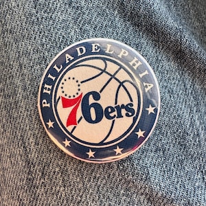 以下が含まれることがあります： 「PHILADELPHIA」と「76ers」の文字が紺色で書かれた丸いボタン。バスケットボールのグラフィックと星が特徴です。背景はデニム生地です。