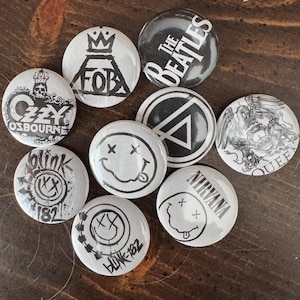 May include: A selection of circular badges with black and white designs, featuring band logos and names. Bands include "The Beatles", "Nirvana", and "Queen". The badges are displayed on a dark wooden surface, showcasing a variety of music-related imagery.