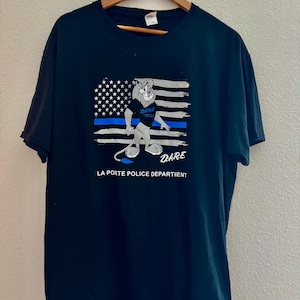 May include: Black t-shirt with a graphic of a cartoon lion wearing a police hat and standing in front of an American flag with a blue line. The text "DARE" is on the lion's chest and below the graphic. The text "LA PORTE POLICE DEPARTMENT" is below the graphic.