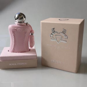 May include: A pink perfume bottle with a silver cap and tassel, presented on a small box. The box next to it is light pink with the words "DELINA" and "PARFUMS de MARLY PARIS" on it. The base of the bottle reads "ROYAL ESSENCE".