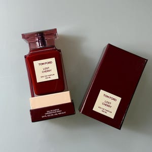 May include: A red-tinted glass perfume bottle and matching box. The bottle and box feature the text "TOM FORD LOST CHERRY EAU DE PARFUM 100 ML". The bottle is rectangular with a square cap. The box is a deep red color.