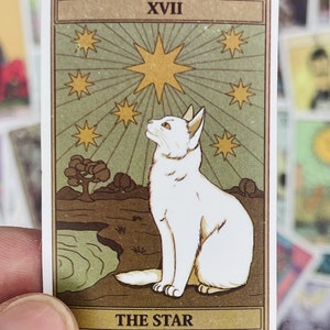 May include: A vintage tarot card with a white cat sitting on a brown rock, looking up at a large yellow star with smaller stars around it. The card is green and brown with the text "XVII THE STAR" at the bottom.