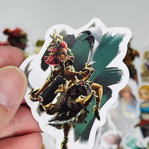May include: A sticker featuring a female character from the Legend of Zelda video game series. The character is depicted in a warrior pose, wielding a sword and wearing golden armour with red accents. The background is a green and black abstract design.