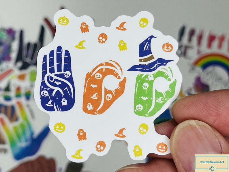 May include: A white sticker featuring three hand gestures spelling out "BOO" in British Sign Language. The hands are decorated with Halloween-themed graphics, including ghosts, pumpkins, bats, and witches' hats.