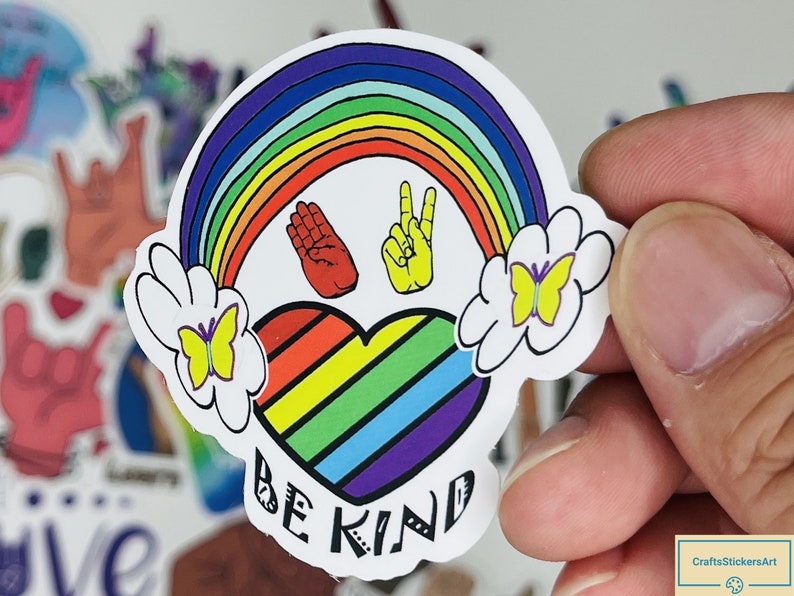 May include: A rainbow-coloured heart with the words "Be Kind" underneath. The heart is surrounded by a rainbow, two clouds with butterflies, and two hand signs for "love" and "peace".