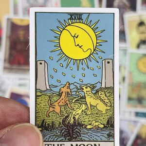 May include: The Moon tarot card, featuring a yellow moon with a face, two towers, two dogs, and a scorpion. The card is blue with a green border and the text "THE MOON."