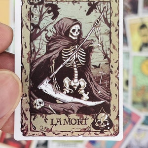 May include: A vintage-style tarot card featuring Death, the 13th card in the Major Arcana. The card depicts a skeletal figure in a hooded robe, holding a scythe and standing in a graveyard. The card is labeled "La Mort" in a decorative font.