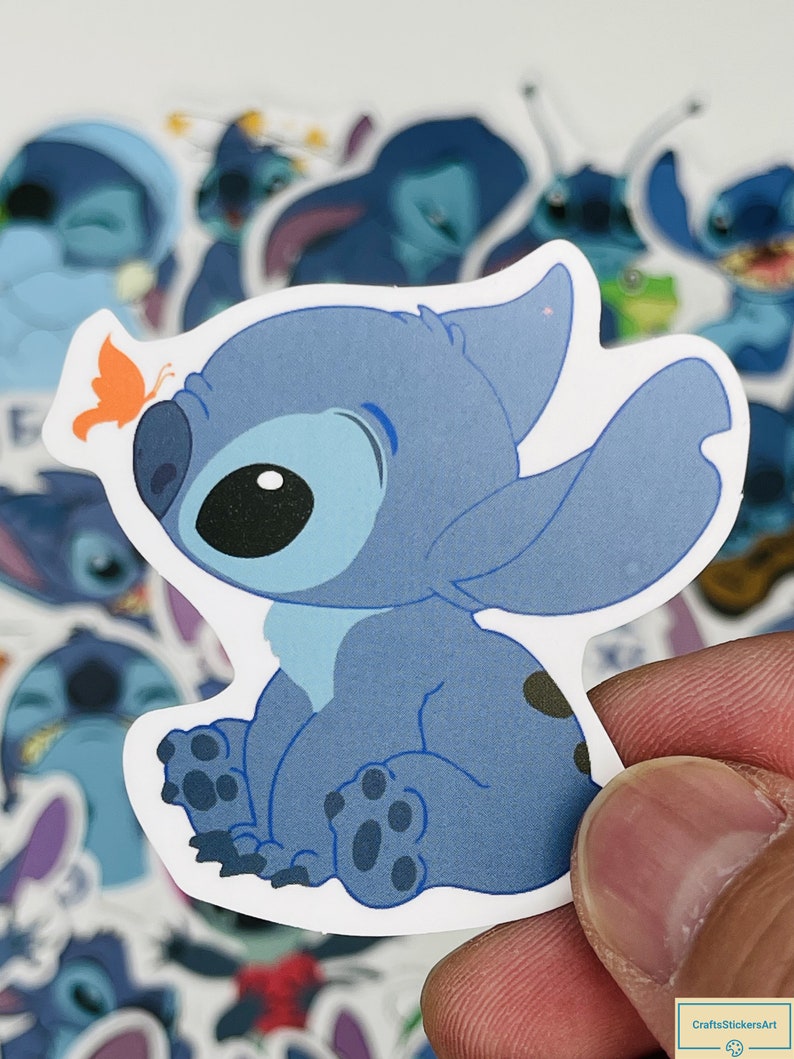 Puede incluir: Una pegatina azul del personaje Stitch con una mariposa en la cabeza. La pegatina est&aacute; sobre un fondo blanco.