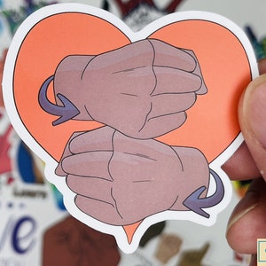May include: A heart-shaped sticker with two hands forming the British Sign Language symbol for "love". The hands are brown and the heart is orange.