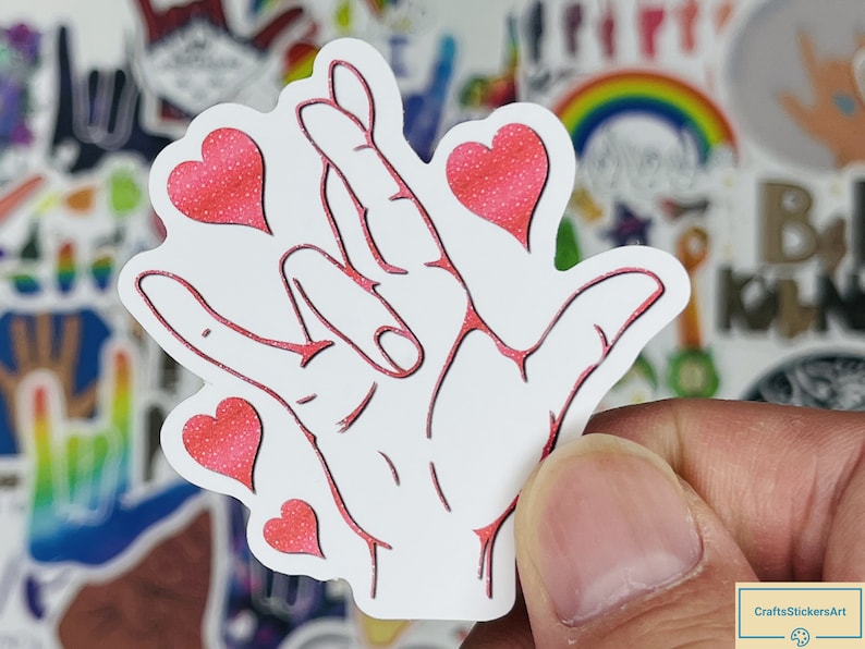 May include: White sticker with a red outline of a hand making the American Sign Language sign for 'I love you'. The hand is surrounded by five red glitter hearts.